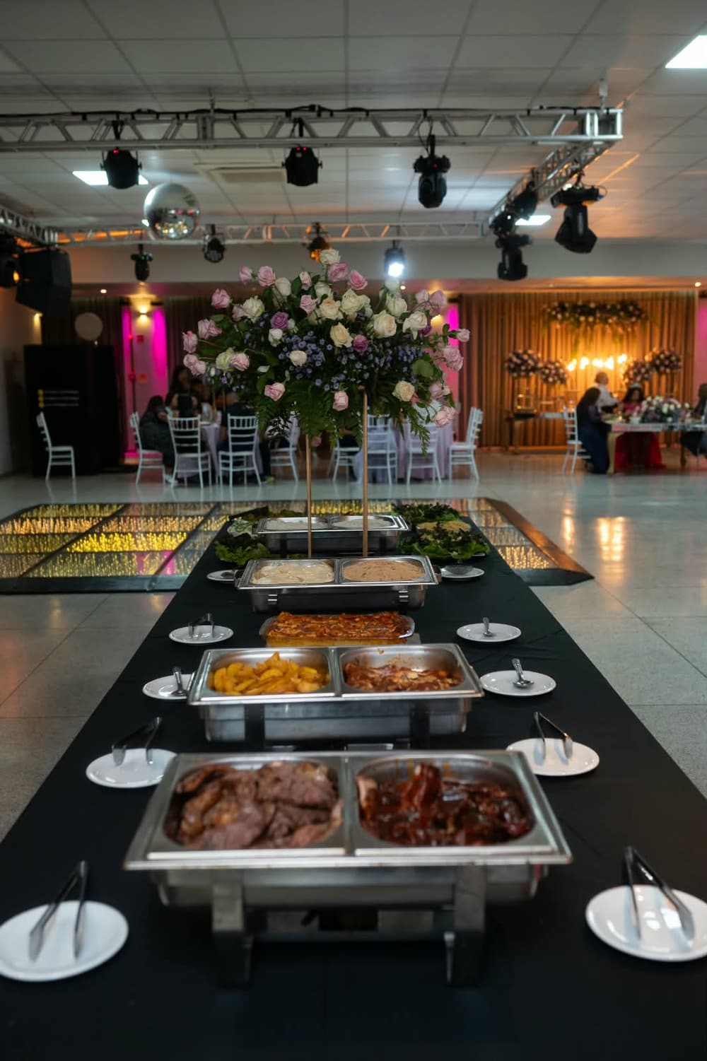 Buffet montado no salão decorado, com arranjos de flores ao fundo
